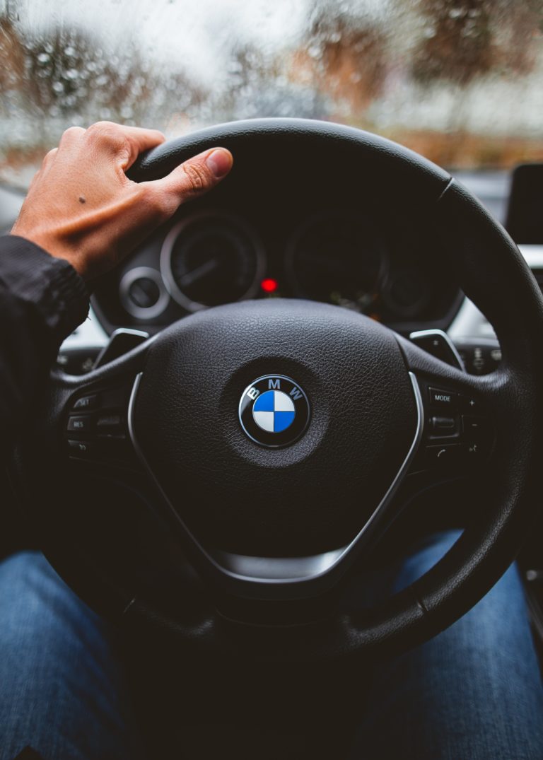 A person’s left hand is holding the steering wheel of a BMW car, with the BMW logo clearly visible, perhaps after choosing a new car. The scene outside the windscreen is blurred, suggesting rainy weather.