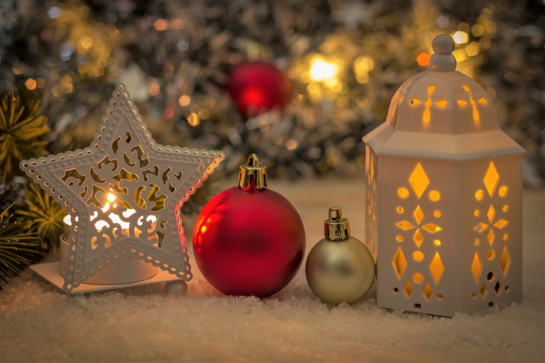 A festive scene with a lit white lantern, a decorative white star, two red and gold Christmas baubles, and blurred lights in the background captures the warmth of Christmas preparation on a snowy surface.