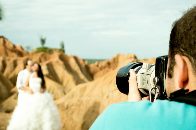 A photographer in a blue shirt takes a picture of a couple posing in front of rocky, desert-like terrain. As they celebrate their big day, the couple—perhaps after delegating wedding duties—stand together, the woman in a white dress.