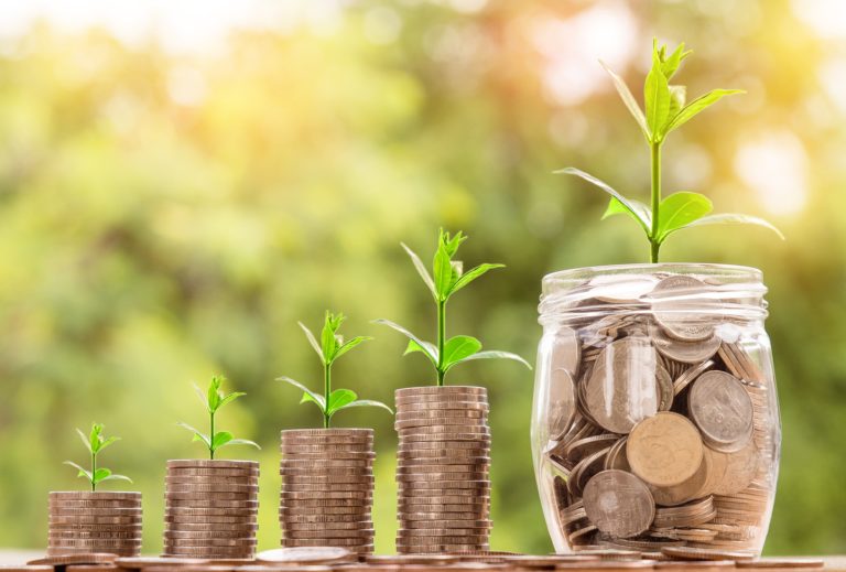Four stacks of coins, each with a small plant growing on top, increase in height from left to right, next to a glass jar full of coins with a larger plant, symbolising smart finances and steady financial growth.