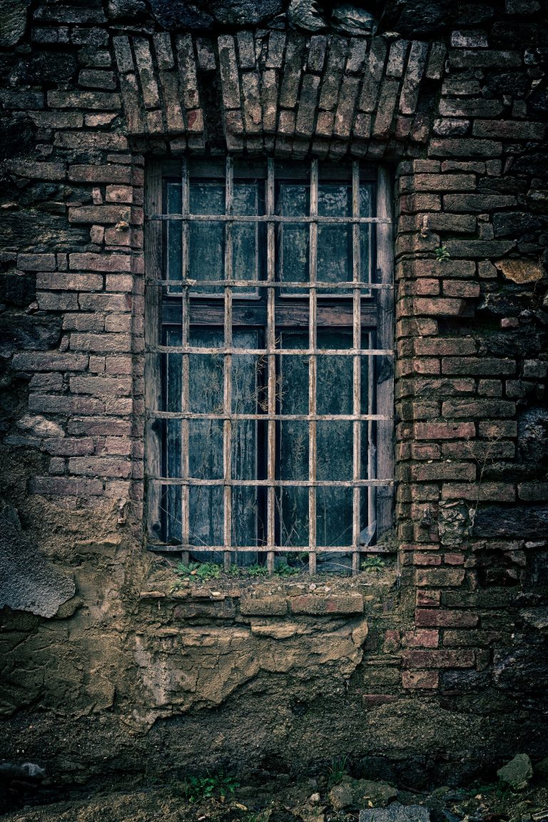 A weathered brick wall with a barred, arched window and a rusted Juliet balcony. The frame is old, and the bricks are chipped and discoloured, giving the scene an abandoned, aged appearance.