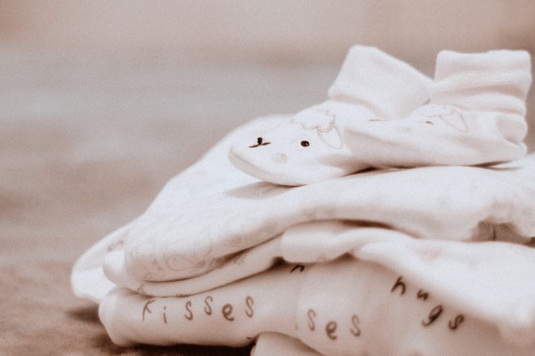 A close-up of folded baby clothes, including a pair of socks with a cute animal face, perfect for a baby on a budget. The clothing features soft colours, delicate patterns, and the words "hugs" and "kisses" visible.