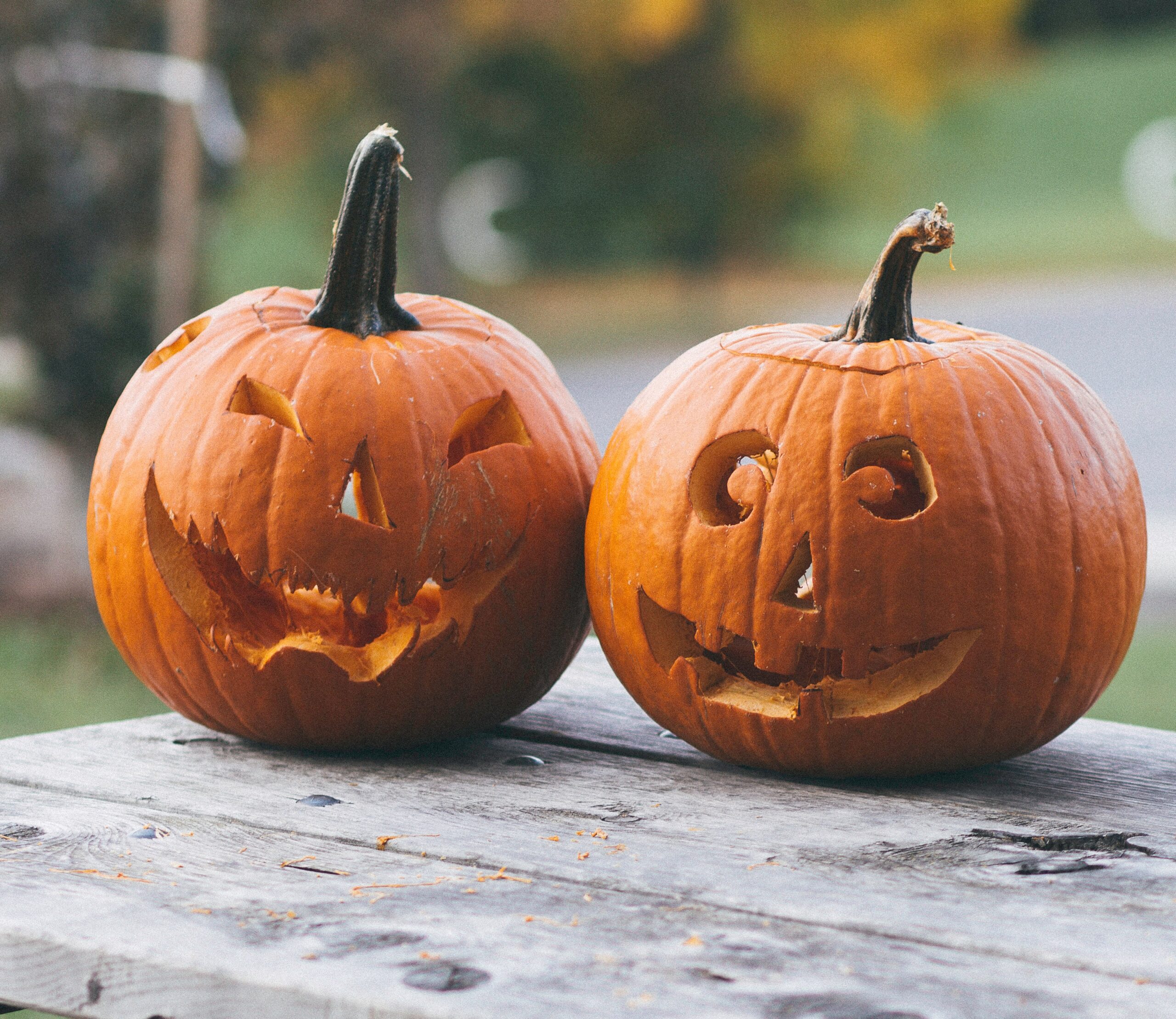 Two carved jack-o’-lantern pumpkins with different faces sit on a weathered wooden table outdoors, with a blurred background of grass and trees. Enter for your chance to win an Amazon voucher this festive season!.