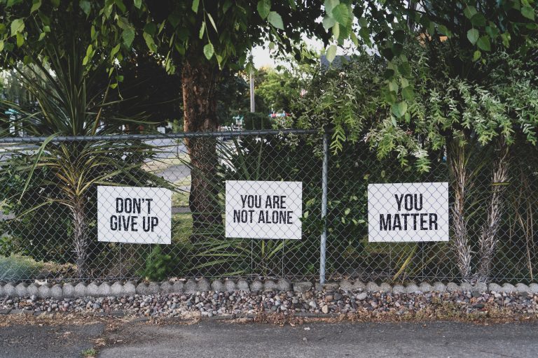 Three signs with encouraging messages—“DON’T GIVE UP,” “YOU ARE NOT ALONE,” and “YOU MATTER”—are attached to a chain-link fence, offering hope for those facing addiction, with green trees and plants in the background.
