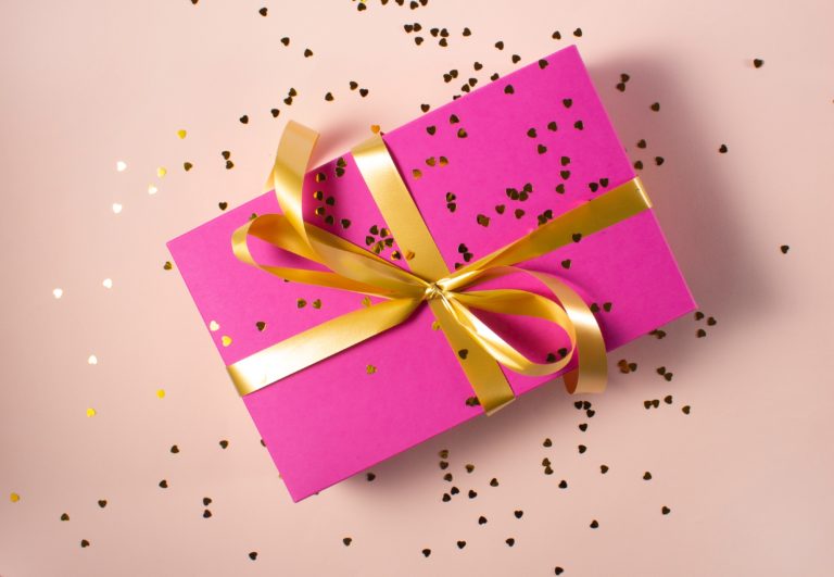 A pink gift box wrapped with a shiny gold ribbon and bow sits on a light pink surface, surrounded by scattered small gold heart-shaped confetti—perfect for any special occasion from Buyagift.
