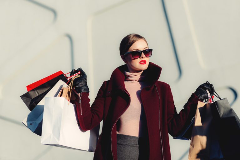 A stylish woman wearing sunglasses, a burgundy coat, and black gloves holds several shopping bags from designer brands in both hands. She stands outdoors against a light, modern wall.