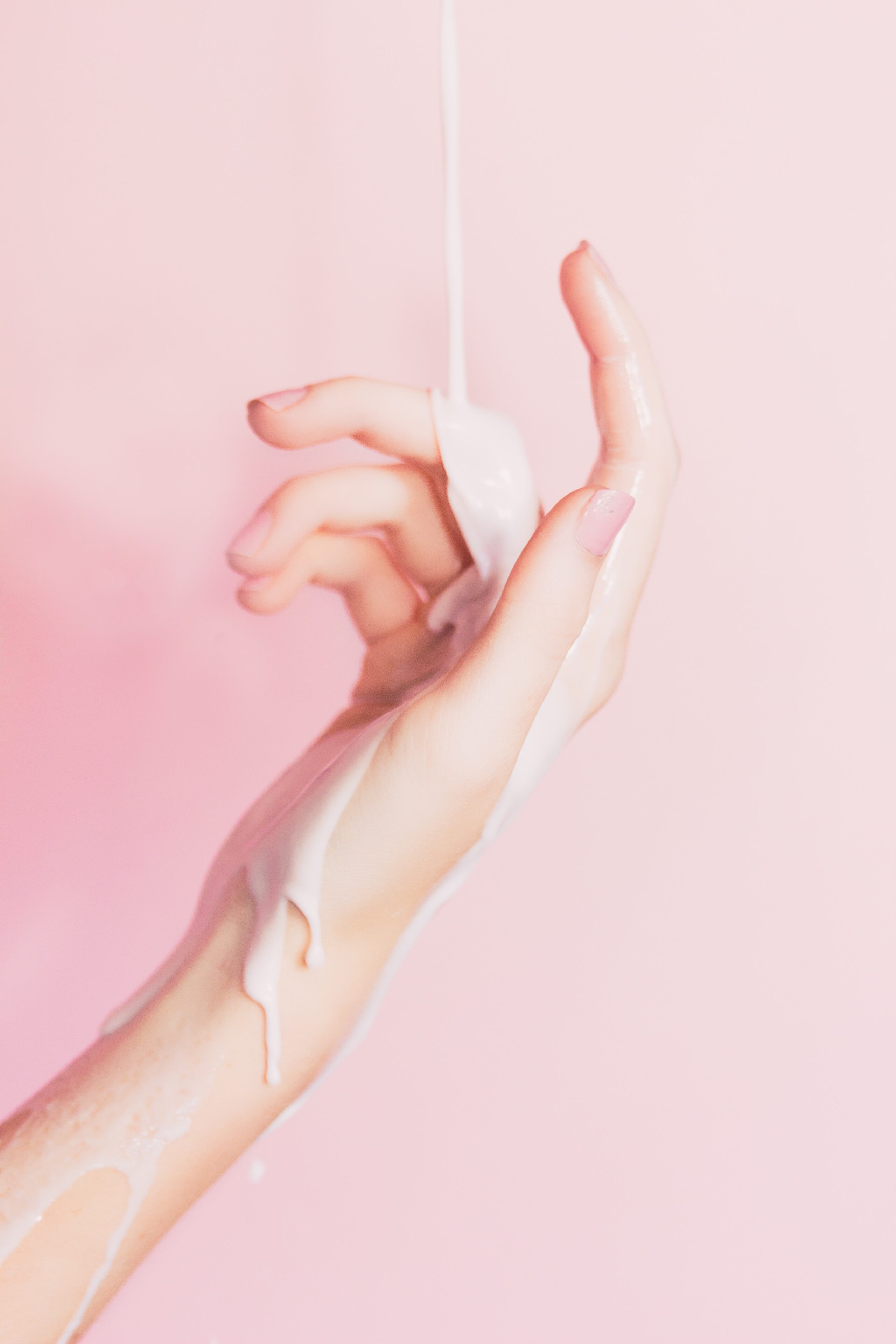 A delicate hand with light skin is posed against a pink background, as a stream of white, fermented skincare liquid pours over the fingers and wrist, creating a smooth, flowing texture.