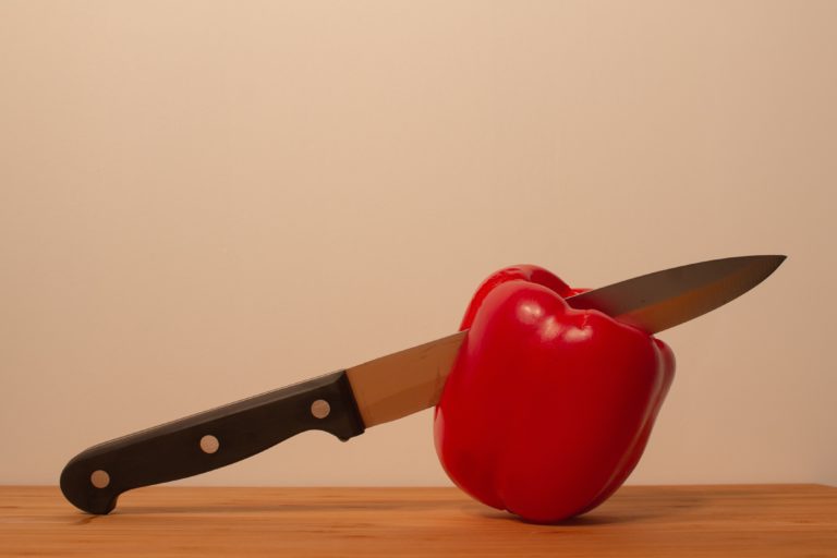 A kitchen knife is partially inserted into a whole red pepper, which is resting on a wooden surface against a plain beige background, highlighting the importance of kitchen safety while preparing fresh ingredients.