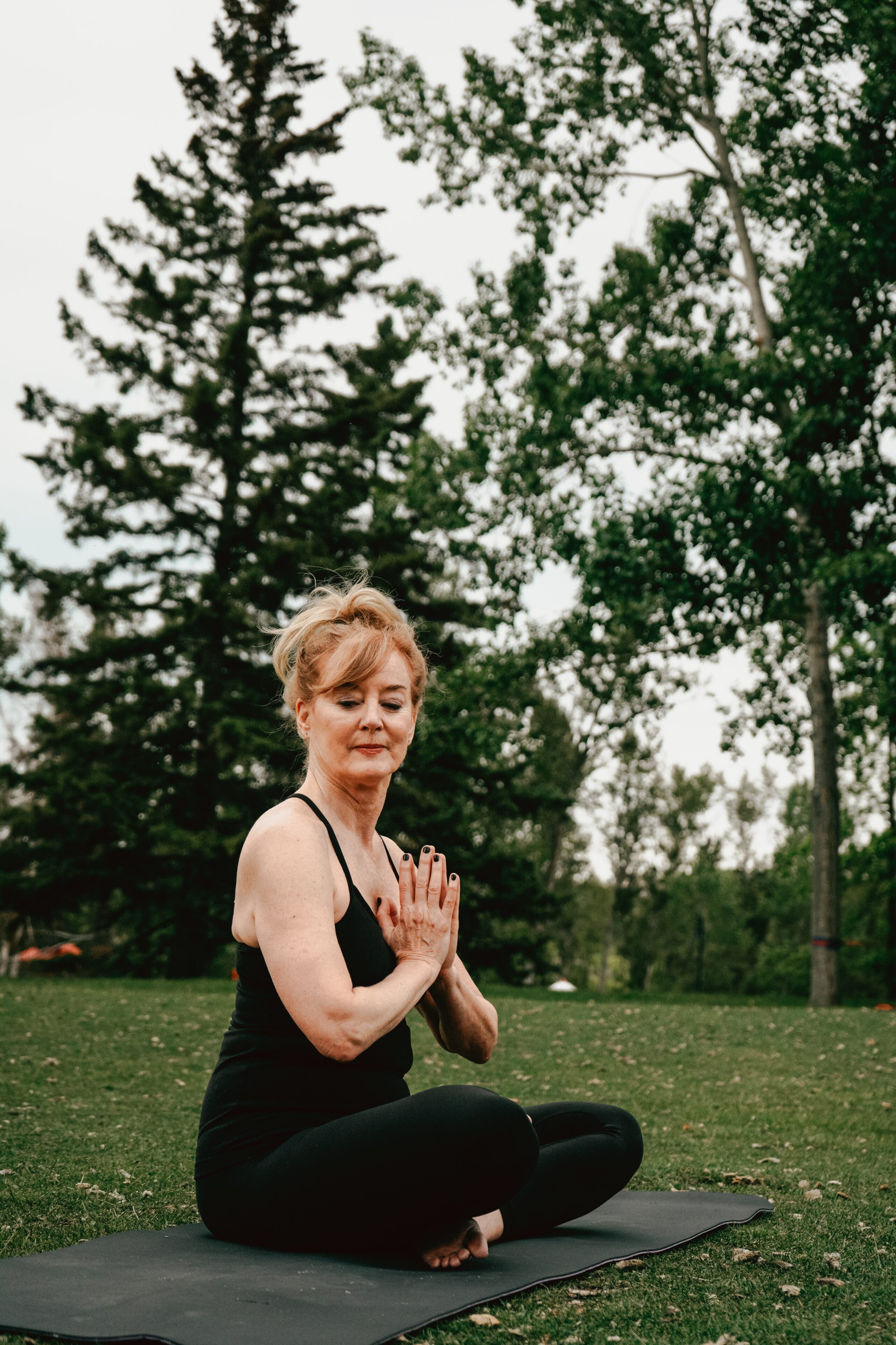 A woman sits cross-legged on a yoga mat outdoors, wearing black workout clothes. Practising meditation—one of the ways to look and feel younger—she holds her palms at her chest, eyes closed, with tall trees in the background.