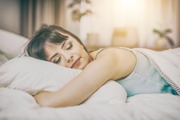 A woman with short brown hair lies on her side in bed, peacefully enjoying a restful sleep on a white pillow and sheet, as soft natural light illuminates the cosy bedroom.
