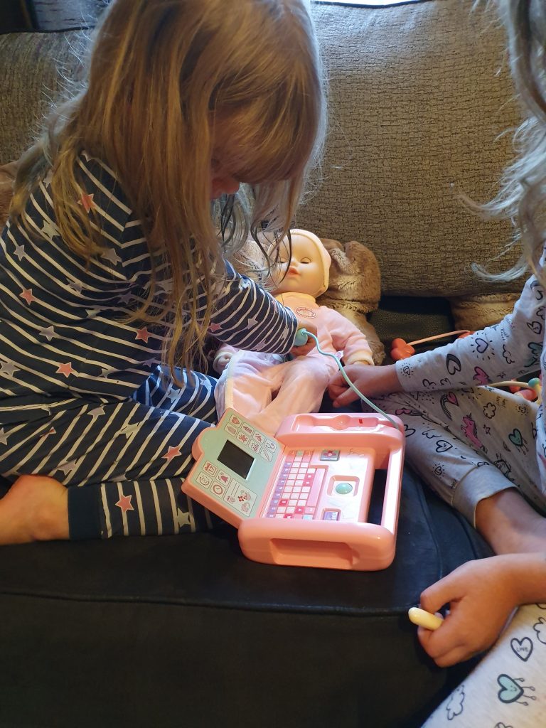 Two young children in pyjamas play doctor with a baby doll, using a toy medical kit that includes a pretend monitor and stethoscope, happily declaring, "Milly feels better," as they sit together on the sofa.