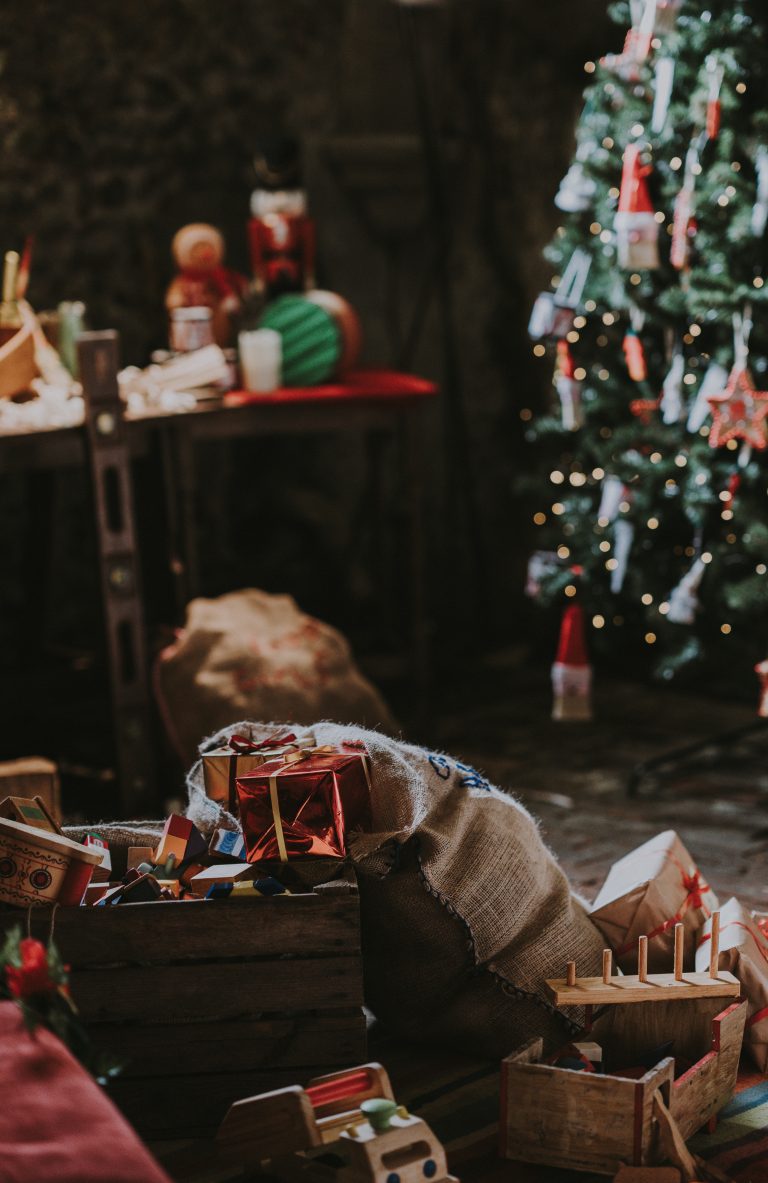 A festive room with a decorated Christmas tree, presents, and wooden toys scattered in crates and hessian sacks, creating a cosy holiday atmosphere filled with charming gift ideas for children.
