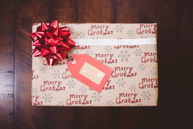 A Christmas present wrapped in brown paper with “Merry Christmas” text, decorated with a large red bow and a red gift tag, sits on a wooden surface—perfect for saving money this Christmas whilst still giving from the heart.