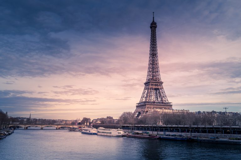 The Eiffel Tower stands beside the Seine River in Paris at sunset, with soft pink and blue tones in the sky and boats moored along the riverbank—a perfect scene for Continental Travel Inspiration for Summer.