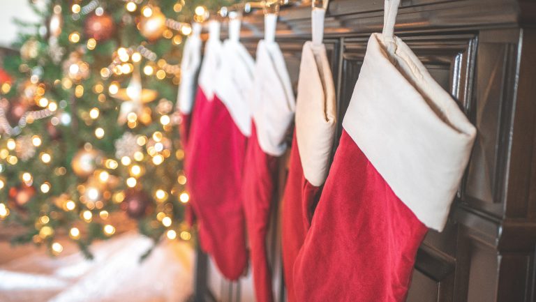 Four red and white Christmas stockings hang from a mantelpiece, ready for stocking fillers, with a decorated and lit Christmas tree blurred in the background. The scene feels festive and cosy.