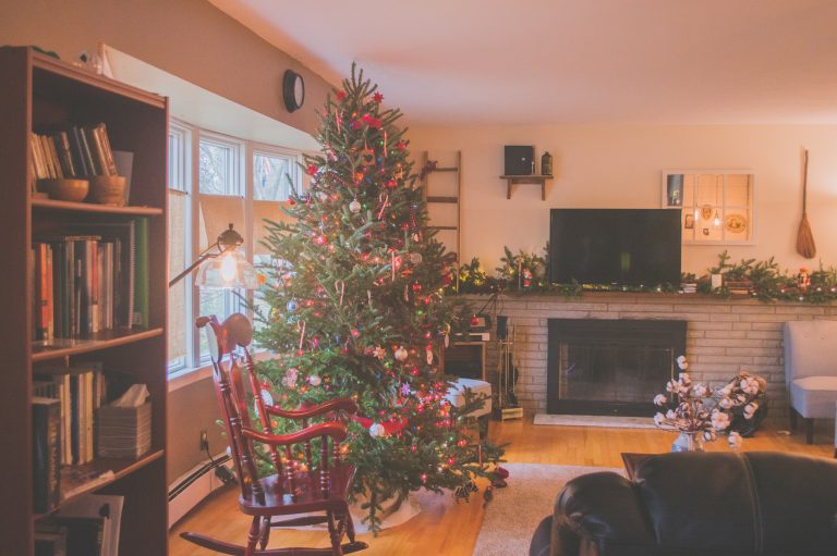 A cosy living room decorated for Christmas features beautiful flooring, a large, decorated tree near the window, a rocking chair, bookshelves, TV above the fireplace, and garlands on the mantelpiece. Warm natural light fills the space.