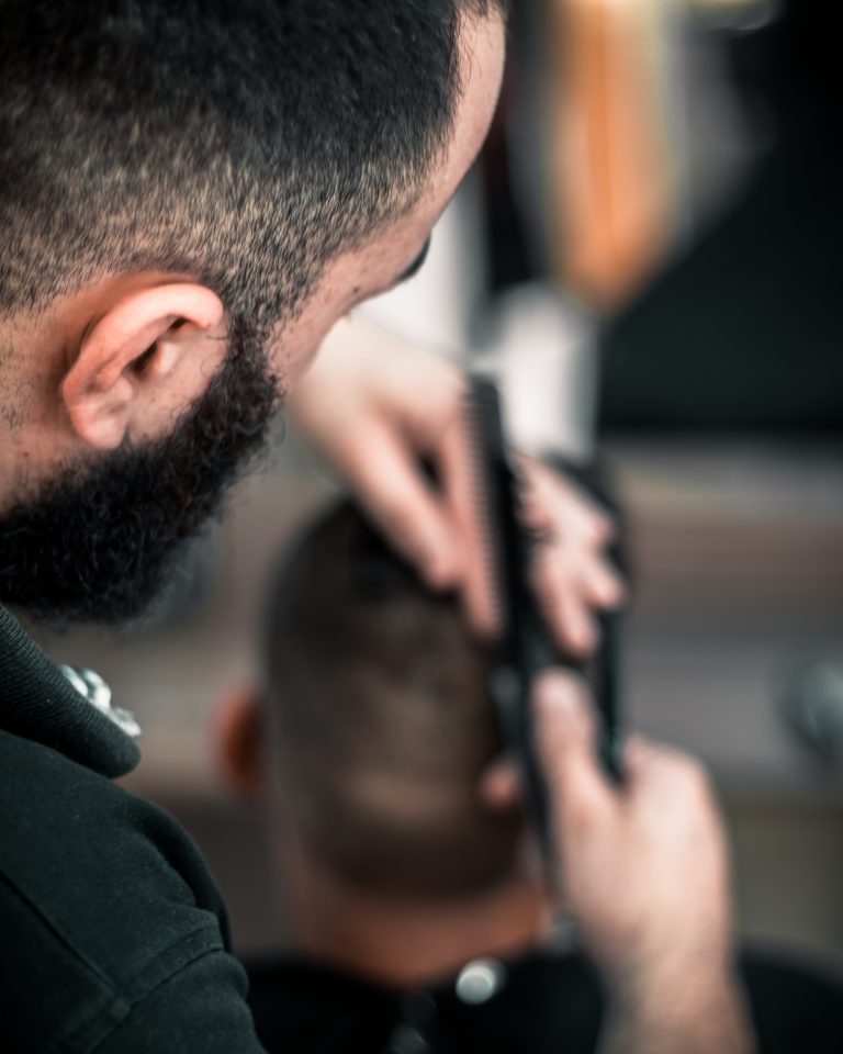 A close-up of a barber with a beard cutting a customer's hair using scissors and a comb. The focus is on the barber's hands and the back of the customer's head, subtly highlighting expert care for hair loss in men. The background is blurred.