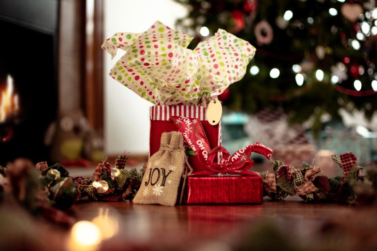 Red and brown Christmas presents with festive ribbons and tissue paper sit by a decorated tree, with a small hessian bag labelled "JOY" and warm lights glowing in the background.