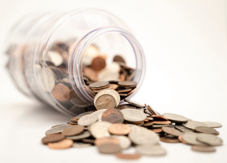 A clear jar tipped over on its side with assorted coins spilling out onto a white surface, symbolising saving money.