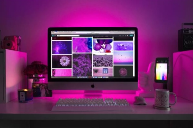 A desktop computer displays purple-themed images on the screen, illuminated by pink-purple LED lighting. A mobile phone open to dating sites for married people, coffee mug, keyboard, mouse, and candle sit on the white desk—a cosy and modern scene.