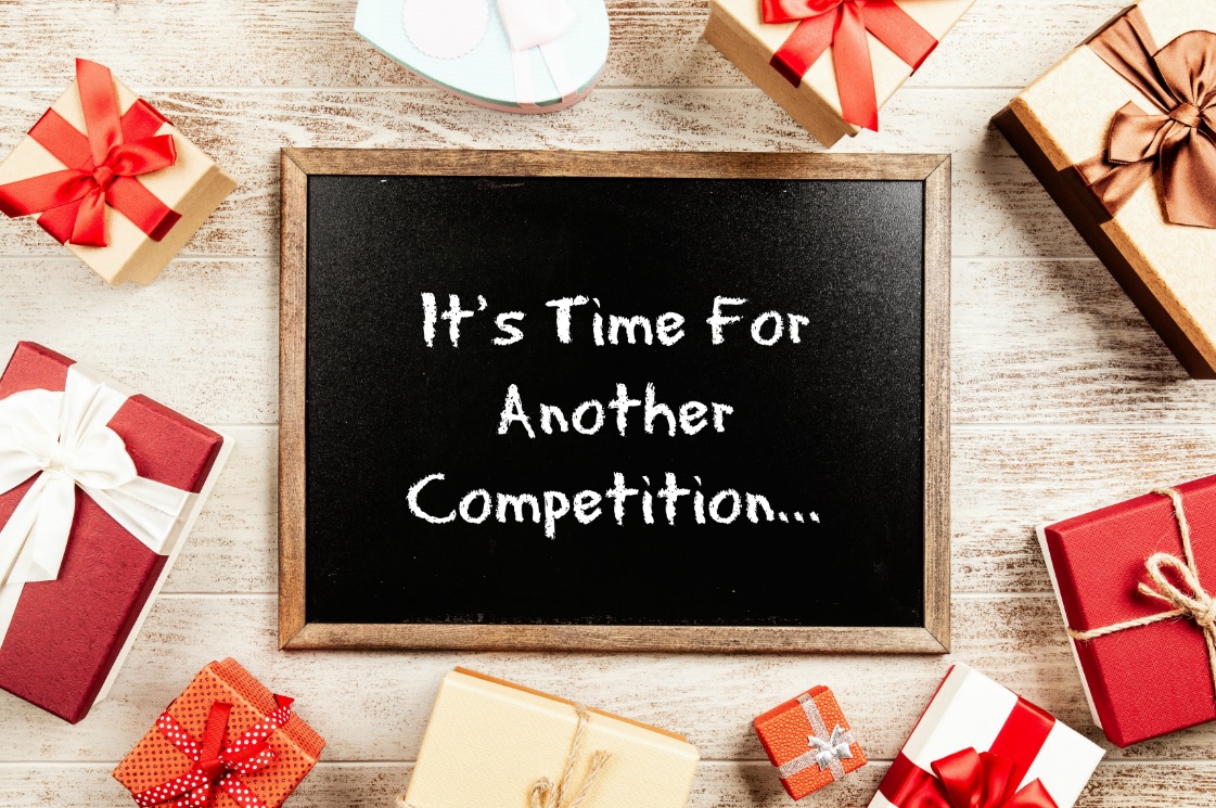 A blackboard with the message "It's Time For Another Competition..." is surrounded by several gift boxes, including the charming Peter Rabbit Year Ahead Bundle, all wrapped in colourful paper and ribbons on a wooden surface.