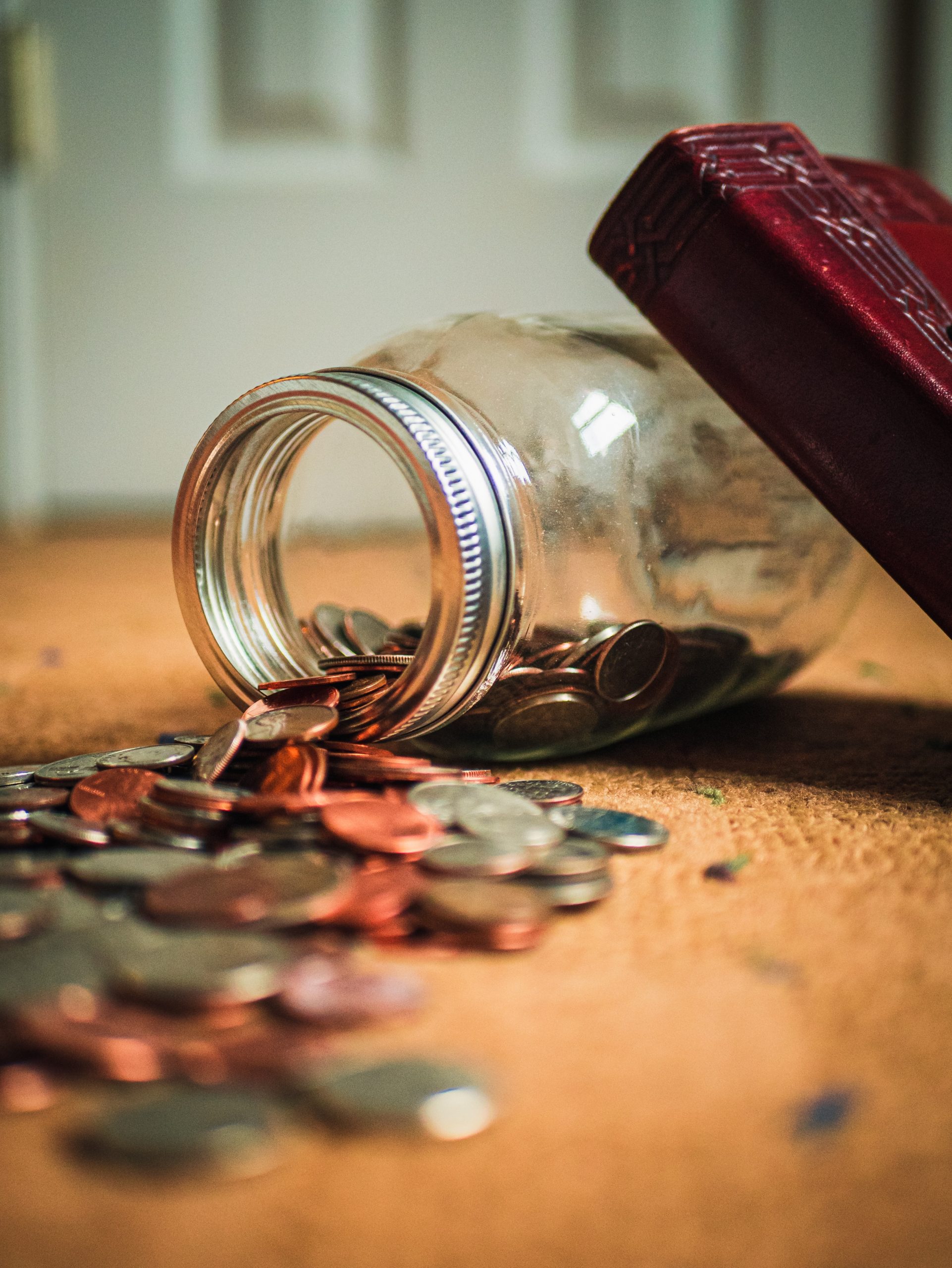A glass jar tipped over on a carpeted floor, spilling assorted coins—a simple scene evoking the importance of teaching finance in schools. Part of a red book rests against the jar, all softly lit for a warm, homely atmosphere.