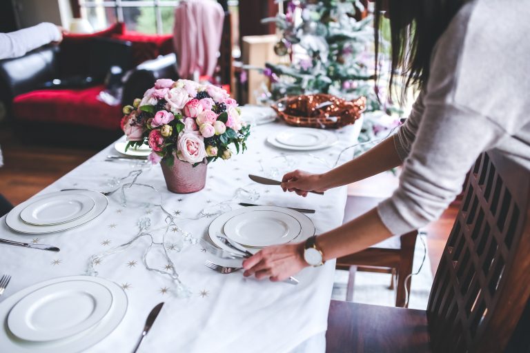A person lays a dining table decorated with a bouquet of pink flowers, plates, and cutlery—perfect for hosting your first dinner party. A Christmas tree and festive decorations are visible in the background.