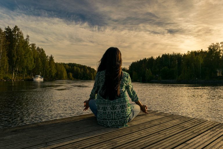 A person sits cross-legged on a wooden jetty by a calm lake, meditating at sunset, surrounded by trees under a partly cloudy sky—embracing 2020, the Year of Mindfulness.