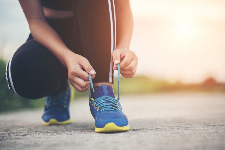 A person in sportswear kneels on a paved surface, tying the blue and yellow laces of their running shoe—highlighting the importance of choosing the right footwear for exercising, with sunlight streaming in the background.
