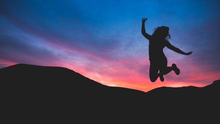 A person is jumping in the air with arms raised, silhouetted against a colourful sunset sky—a vivid reminder of how to find energy to achieve your goals, with pink, purple, and blue hues lighting up the hills in the foreground.