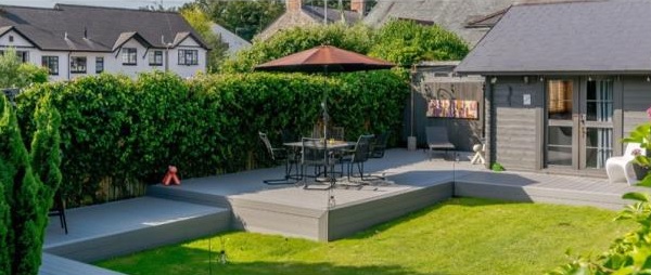 A modern back garden perfect for preparing your garden for spring, with a raised grey deck featuring a round table, chairs, and large umbrella. Green hedges and lawn surround a sleek grey garden shed, with houses visible in the background.