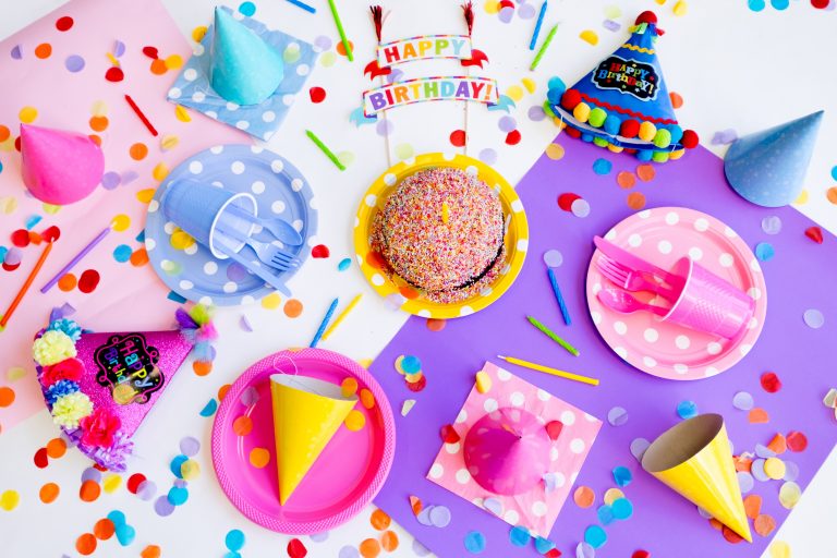 Colourful birthday party scene with confetti, party hats, a sprinkle-covered cake, polka dot plates, cups, forks, and a "Happy Birthday" sign arranged on pink and purple paper backgrounds.