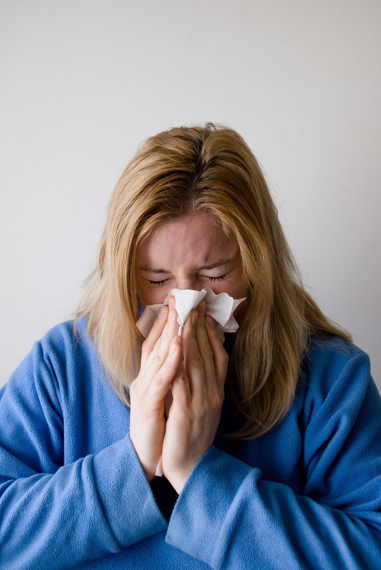 A person with blonde hair wearing a blue dressing gown is sneezing or blowing their nose into a tissue, eyes closed and an expression of discomfort, possibly due to allergies, against a plain light background.