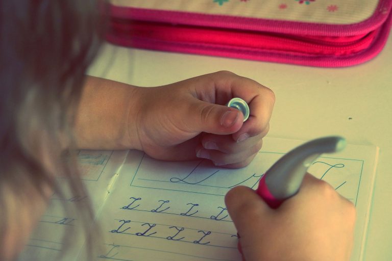 A child practises writing cursive letters in an exercise book, holding a pen in one hand and steadying the paper with the other. A pink pencil case is visible in the background—an engaging way to help your children to succeed.