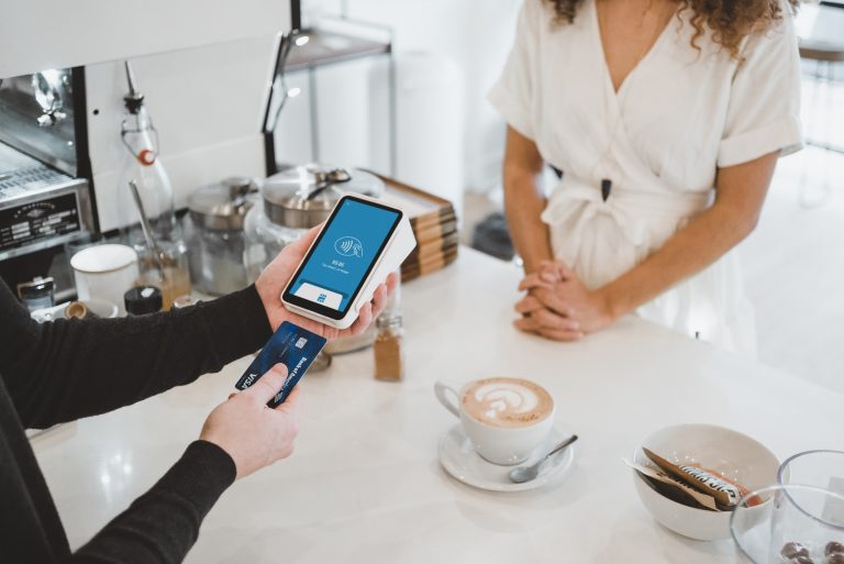 A person makes a contactless payment with a credit card and smartphone at a café counter, while a barista in a white outfit stands opposite them. On the counter are coffee, sugar sachets, and a Corey Advisors flyer.