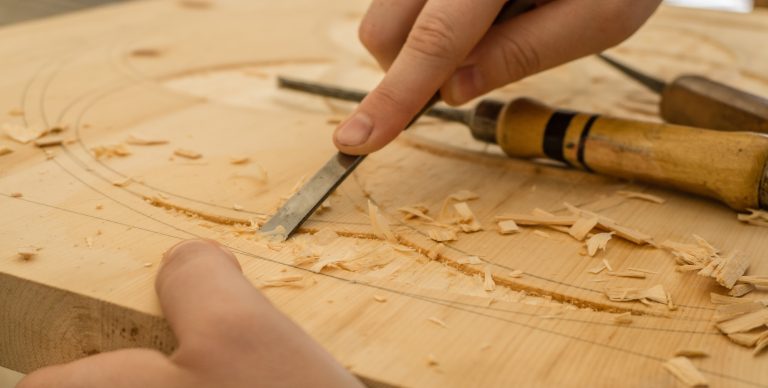 A close-up of hands using a chisel and carving tool to carve designs into light-coloured wood, with wood shavings scattered around—showcasing the craftsmanship behind made-to-measure furniture.