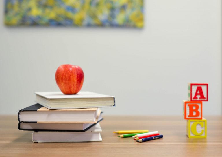 A red apple sat on top of a stack of four books next to a pile of colouring pencils and three building blocks that have the letters ABC on them