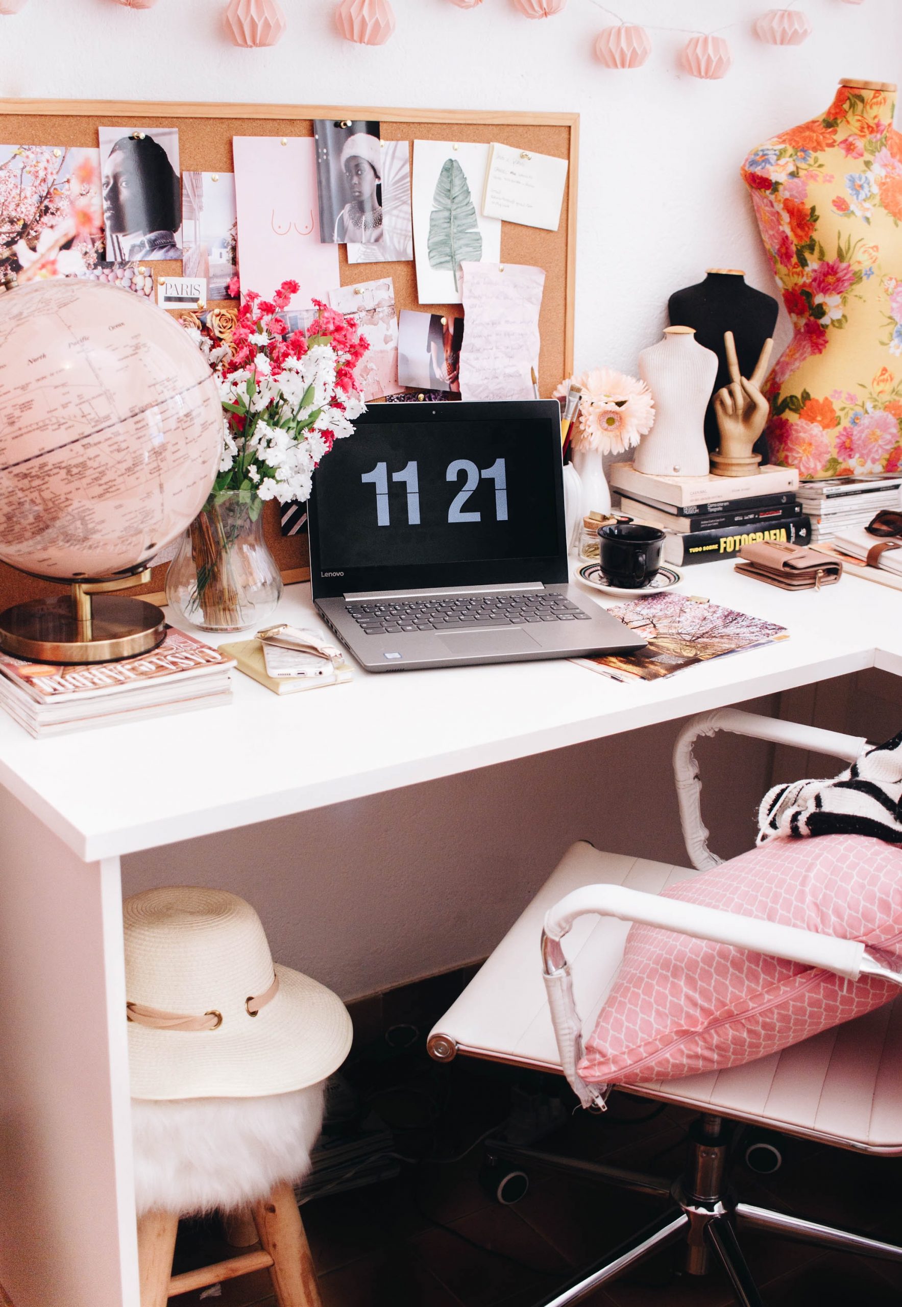 A stylish desk setup reflecting the pros and cons of being self-employed, with a laptop displaying 11:21, books, a globe, flowers, and a camera. Above the desk, a noticeboard holds notes and photos; a white chair and hat await nearby.