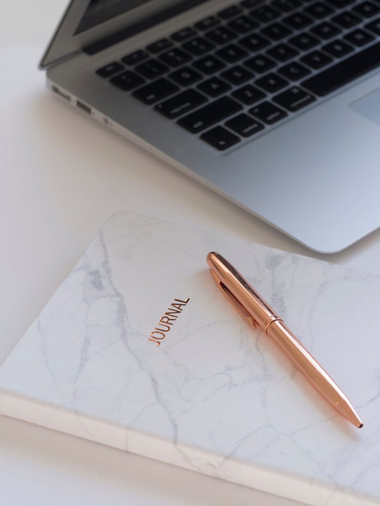 A marble-patterned notebook with "JOURNAL" on the cover and a rose gold pen rests on a white surface next to an open silver laptop—perfect for jotting down ideas about paid work from blogging.
