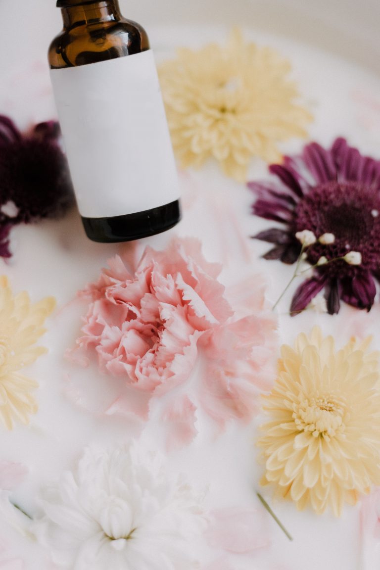 A brown glass dropper bottle labelled for CBD hovers above vibrant pink, purple, yellow, and white flowers floating in milky water, creating a soothing botanical scene.