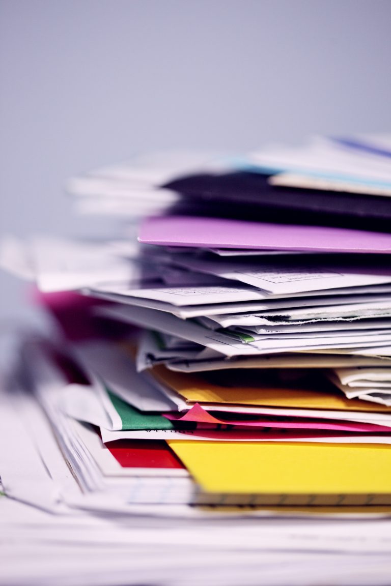A close-up of a messy stack of papers and colourful folders piled on a desk, highlighting the need for office waste management, with a blurred background.