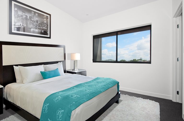 A modern, minimal bedroom features a large bed topped with white bed linen and a teal throw—showcasing reasons to make your next mattress a memory foam mattress for ultimate comfort—plus a bedside lamp, cityscape photo, and window with blue skies.