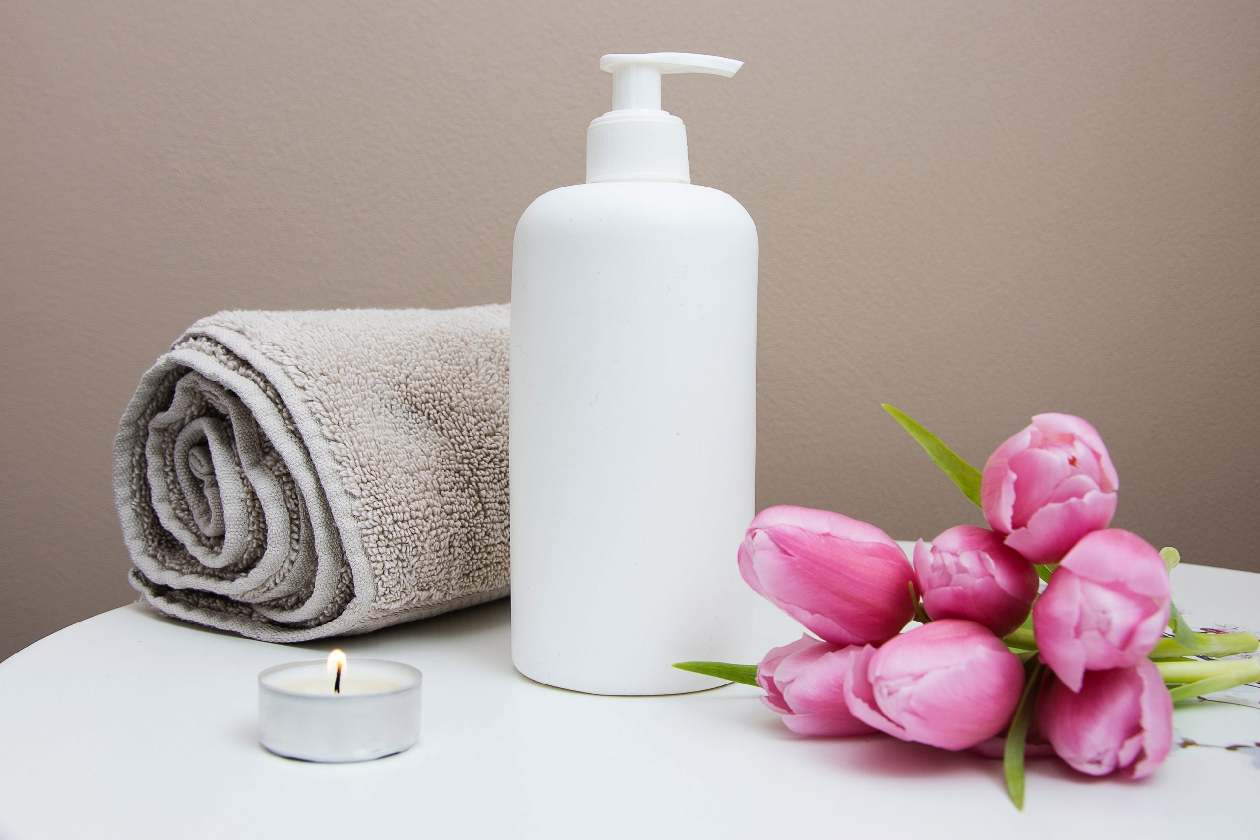a white pump bottle sat next to a rolled up grey towel, a lit tealight candle and a bunch of purple closed tulips