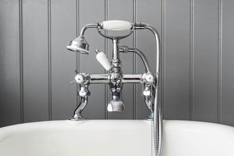 A close-up of a vintage-style chrome bath tap with a handheld shower attachment, mounted on a white bath in front of a grey panelled bathroom wall.