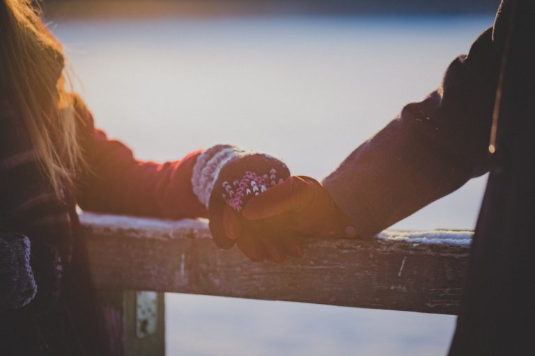 a picture showing the arms of a man and a woman holding hands on a fence in winter as they are both wearing gloves and coats