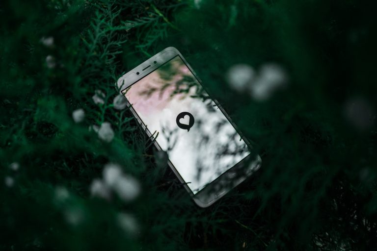 a mobile phone in a green bush. On the screen is a love heart emoji