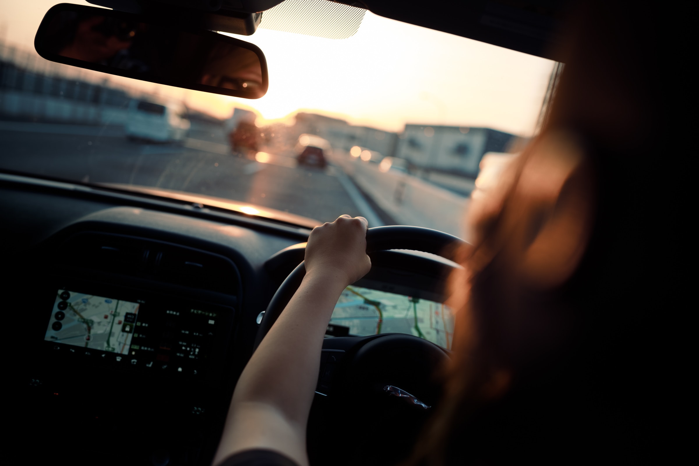 A person drives an affordable family car at sunset, holding the steering wheel with one hand. The dashboard and satnav screen are visible as the sun shines through the windscreen onto the road ahead.