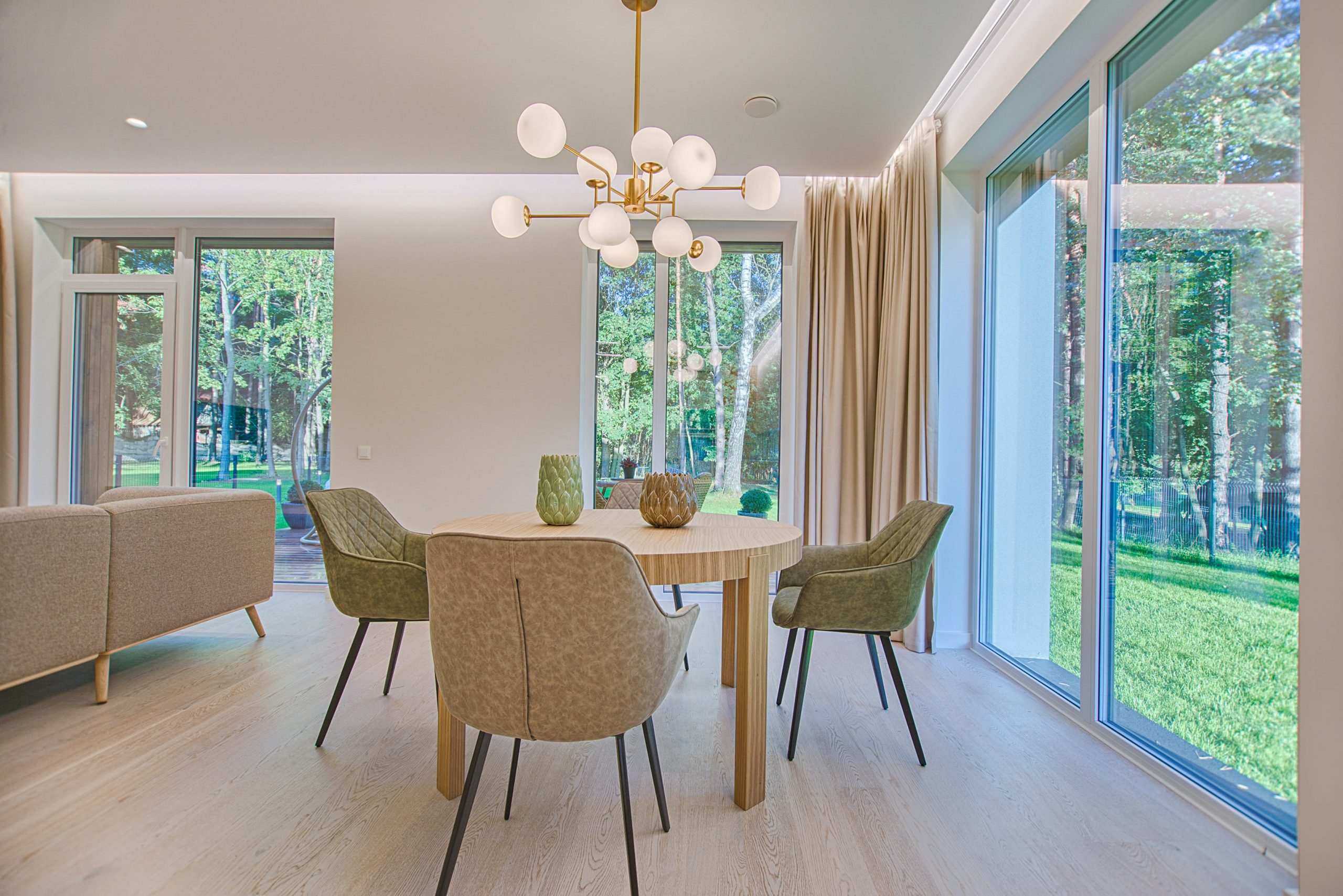 Modern dining area with a round wooden table, four upholstered chairs, and large windows showing a green outdoor landscape. Wooden vinyl flooring adds warmth, whilst a contemporary chandelier hangs above the table, filling the room with natural light.