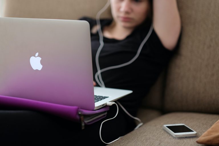 A teenager sat listening to music whilst working on a laptop with a mobile phone sat at the side of her