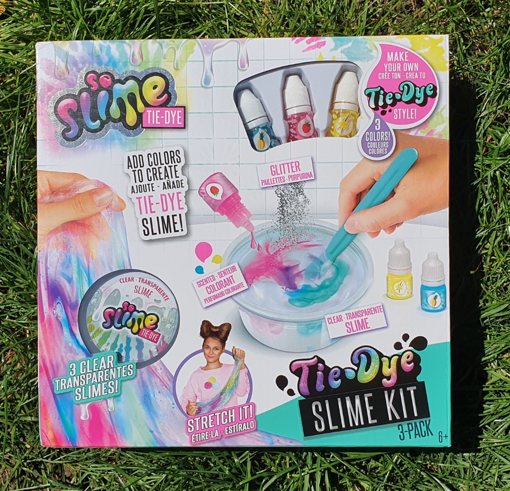 A colourful So Slime Tie-Dye Slime Kit box on grass, showing bottles of dye, a mixing bowl, and hands stirring slime—one of the best toys to keep children occupied with 3 clear slimes and endless colour and glitter customisation.